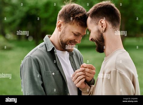 Two Bearded Gay Men Stand Close Together In A Green Park Their Heads Touching As They Share A