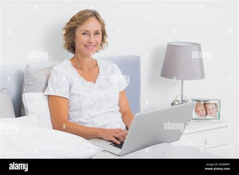 Content Blonde Woman Sitting In Bed Using Laptop Smiling At Camera At Home In Bedroom Stock