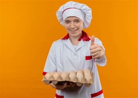 Jeune femme blonde souriante chef en uniforme de chef détient lot d oeufs et pouces vers le haut