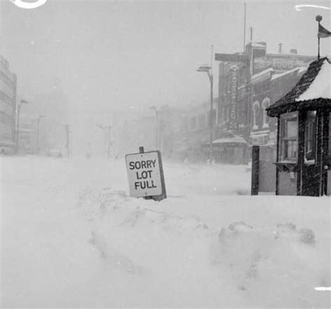 A Sign That Is In The Snow By Some Buildings And People Are Walking