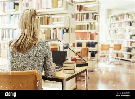 Blonde Female Student Is Sitting At The Desk With Pile Of Books University Library Stock Photo