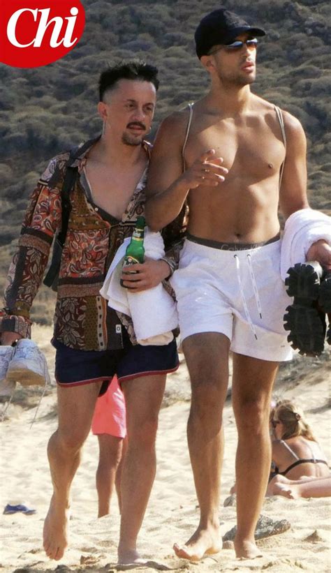 Two Shirtless Men Walking On The Beach With One Holding A Beer And