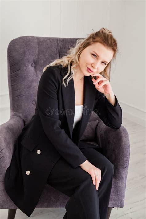 A Cute Blonde Girl In A Black Suit Sits In A Chair And Looks Thoughtfully Into The Camera Stock