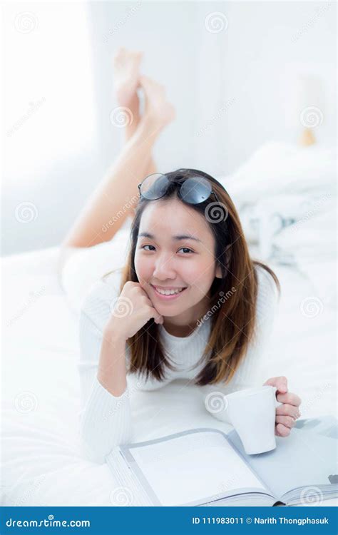 Charming Woman Lying On Bed Reading A Book And Holding Coffee Cup Stock