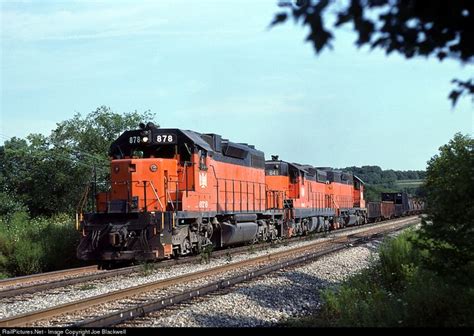 An Orange And Black Train Traveling Down Tracks Next To Some Trees On A