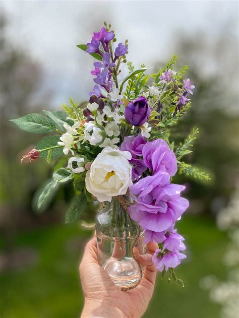 a person holding a vase with purple and white flowers in it's centerpiece