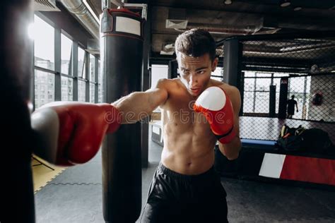 Sportsman In Red Boxing Gloves With A Naked Torso Hits Punching Bag In The Gym On The Background