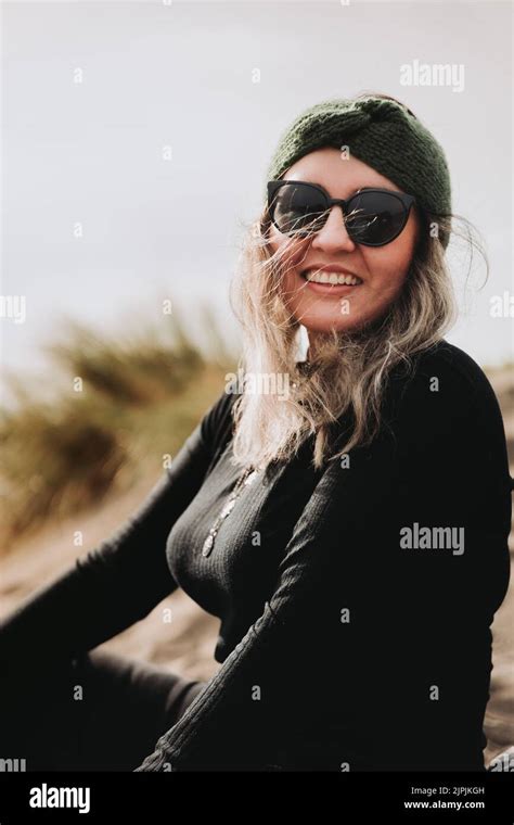 Smiling Blonde Woman Dressed In Black Wearing A Headband Sunglasses And Sitting On The Beach