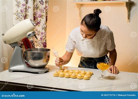 Woman Confectioner Bent Over The Kitchen Table And Squeezes Cream Onto Halves Of Macaroons Stock