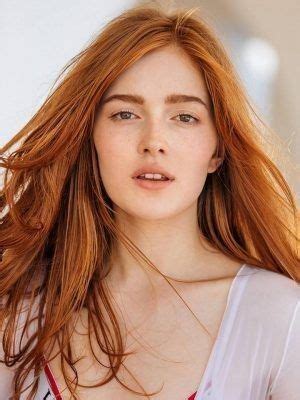 A Woman With Long Red Hair Wearing A White Top And Looking Off To The Side