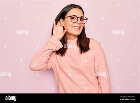 Jeune femme brunette portant un pull décontracté et des lunettes sur fond rose souriant avec