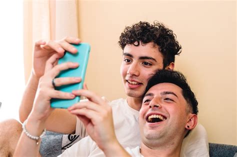 Joven pareja gay sonriente usando el teléfono inteligente para ver contenido entretenido rutina