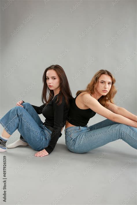 Stylish Photo Shoot Of Two Models Blonde And Brunette On A Gray Background In Jeans And A Black