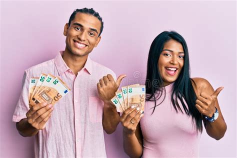 Joven Pareja Latina Sosteniendo Billetes De Euros Apuntando El Pulgar Hacia Un Lado Sonriendo