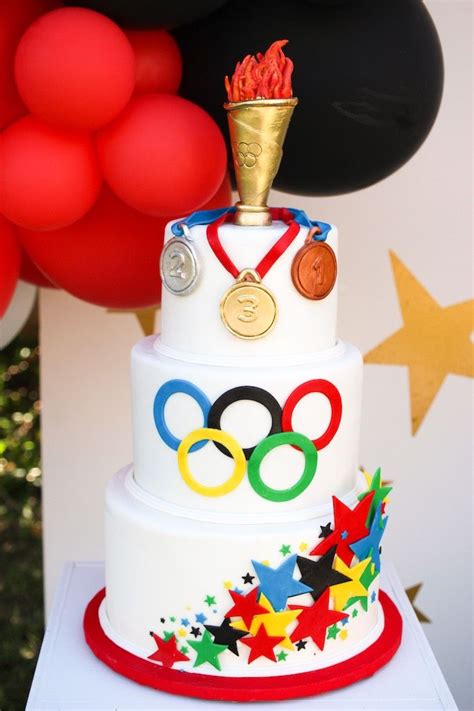 A Three Tiered Cake With Olympic Symbols On It And Red Balloons In The