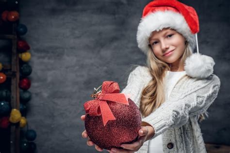 Jolie Fille Blonde Au Chapeau Du Père Noël Tient Une Boule De Noël Rouge Photo Gratuite