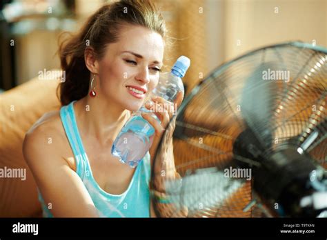 Hot Woman With Bottle Of Cold Water Using Electric Metallic Fan At Modern Home In Sunny Hot