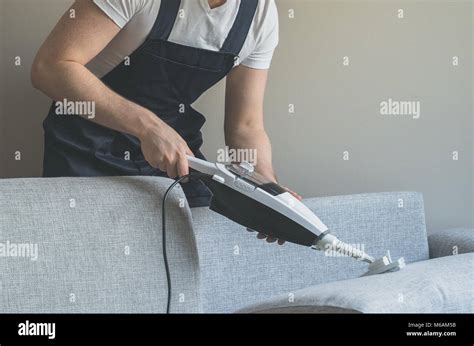 Man In Uniform Cleaning Fabric Of The Sofa With Dry Steam Cleaner Stock