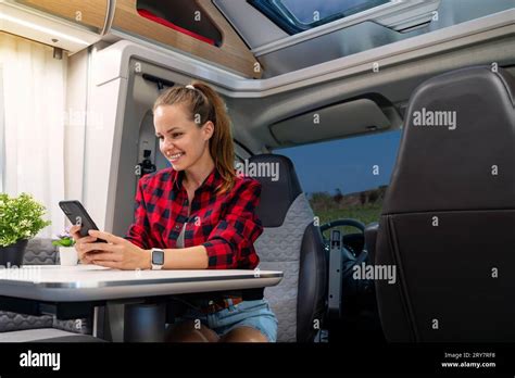 Happy Brunette Woman Sitting At The Table In Camper Van And Using Her Smart Phone RV Rental