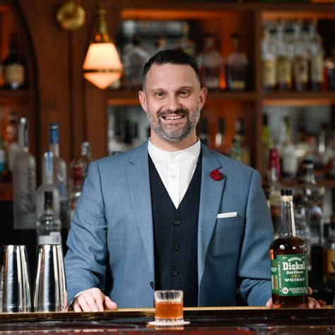 A Man In A Blue Suit Standing Behind A Bar With Bottles And Glasses On It