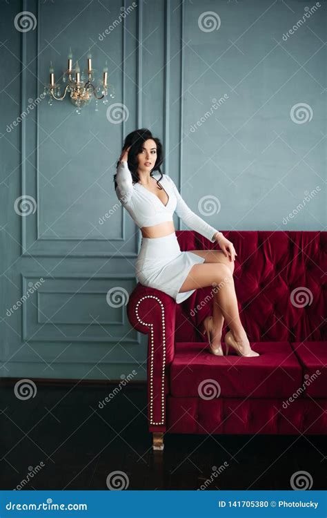 Brunette Girl Wearing In Blue Dress And High Heels Sitting On Burgundy Couch Stock Photo