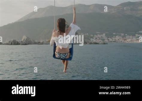 Back View Brunette Woman In A Swimsuit Sits On A Swing Above The Sea Against Mountains Stock