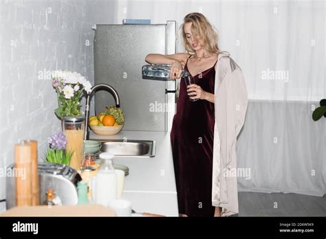 Thirsty Blonde Woman In Trench Coat And Velour Dress Pouring Water Into Glass In Kitchen Stock