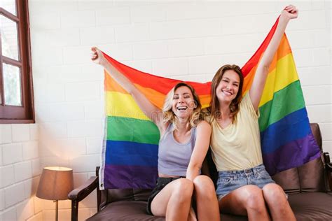 Portraits Of Happy Caucasian Lesbian Couple Sitting And Embracing On A Sofa And Raising Rainbow