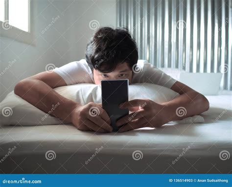 Relaxed Asian Man With Mobile Smart Phone Lying Down On The Bed In