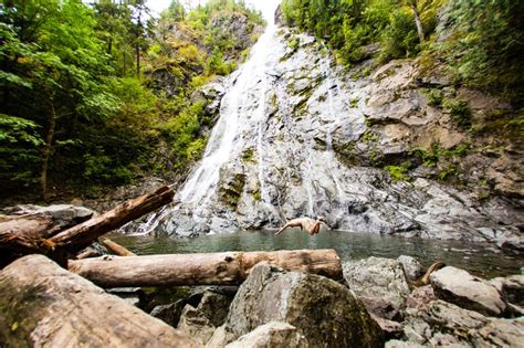 There Is A Man Swimming In The Water Near A Waterfall With Logs And