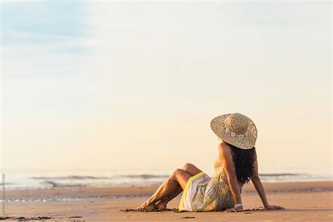 Woman In A Dress With A Sunhat On Sitting On The Beach By Stocksy