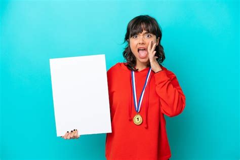 Joven latina con medallas aisladas de fondo azul sosteniendo un cartel vacío y gritando Foto
