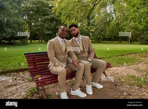 Gay Newlywed Couple Sitting On A Wooden Bench Holding Hands In A Natural Environment They Are