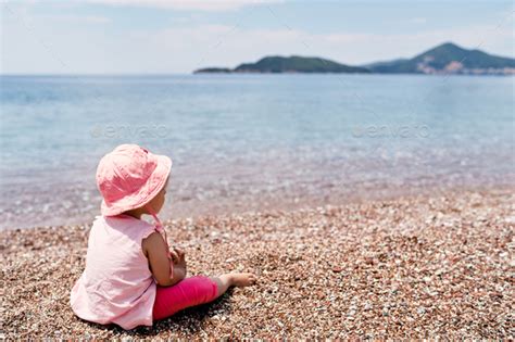 Little Girl In A Panama Hat Sits On A Pebble Beach Near The Water Back