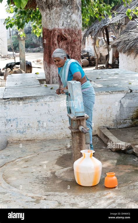 Elderly Indian Woman Pumping Water From A Hand Pump Into A Pot In A