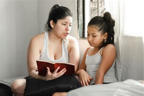 Madre latina enseñando a su hija pequeña la importancia de leer un libro sentada encima de la