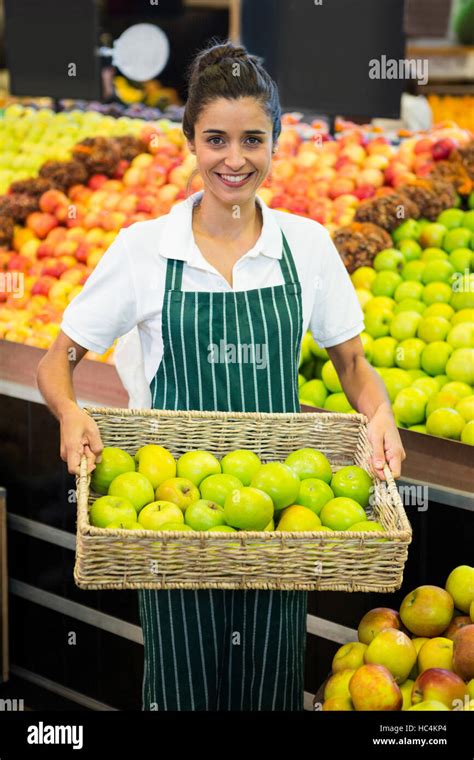 Smiling Female Staff Holding A Basket Of Green Apple At Supermarket