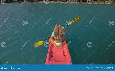 Seductive Blonde Woman In Blue Swimsuit Sails On Pink Plastic Kayak Along Azure Sea At Exotic