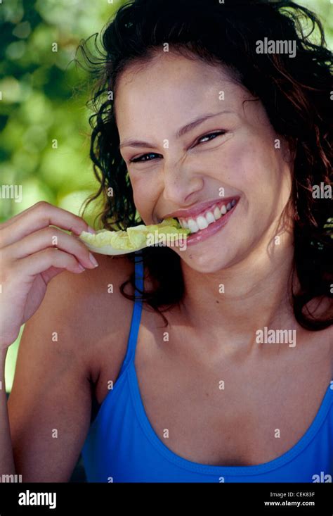 Female Long Curly Brunette Hair Wearing Blue Vest Top Eating Lettuce Leaf Looking To Camera