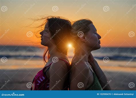 Blonde And Brunette Girlfriends Are Clinging Back To Each Other Against Sunset Over The Sea