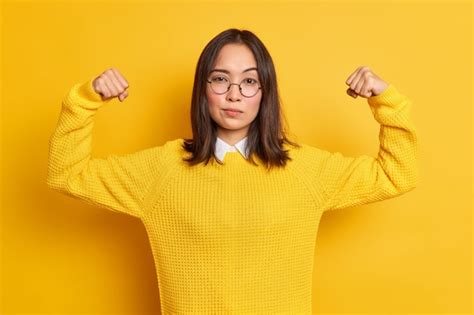 자신감이 넘치는 젊은 아시아 여성은 영웅이 그녀의 힘을 보여주고 힘을 보여주는 것처럼 팔 근육이 둥근 광학 안경 스웨터를