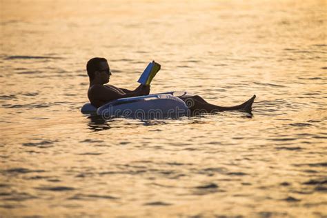 Caucasian Man Reads A Book Floating On The Sunset In The Ocean Water