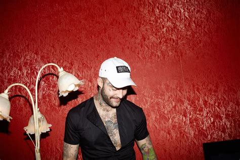 A Man With Tattoos Standing In Front Of A Red Wall Wearing A White Hat