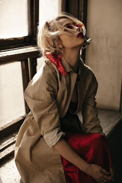 Charmante dame bouclée blonde en trench coat beige lunettes de soleil rouges et jupe en soie