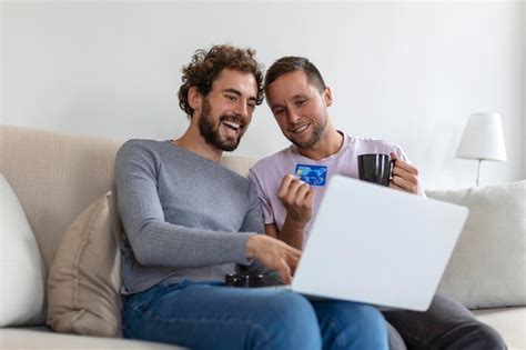 Alegre joven pareja gay sonriendo alegremente mientras compra en línea en casa dos jóvenes