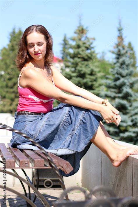 Young Girl In Denim Skirt Sitting Barefoot On A Bench Stock Photo