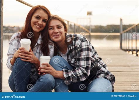 Cute Lesbian Couple Sits Together Near The Lake With Cups Of Drink In Hands And Embracing Each