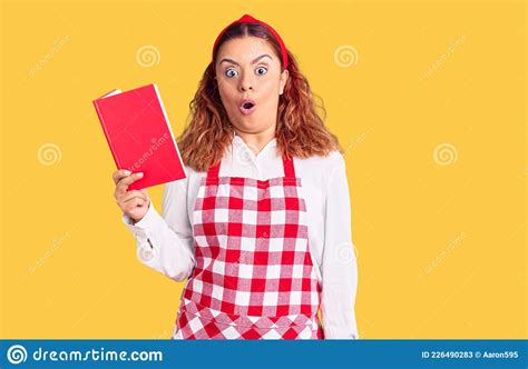 Jovem Latina Vestindo Avental Segurando O Livro De Receitas Assustada E Surpreendida Boca
