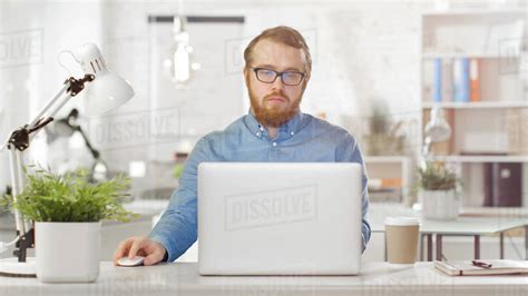 Portrait Of A Young Bearded Man With Glasses Sitting At His Desk Using