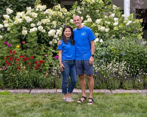 Middle Aged Caucasian Man And His Korean Wife Posing In Front Of A Bed Of Beautiful Flowers In